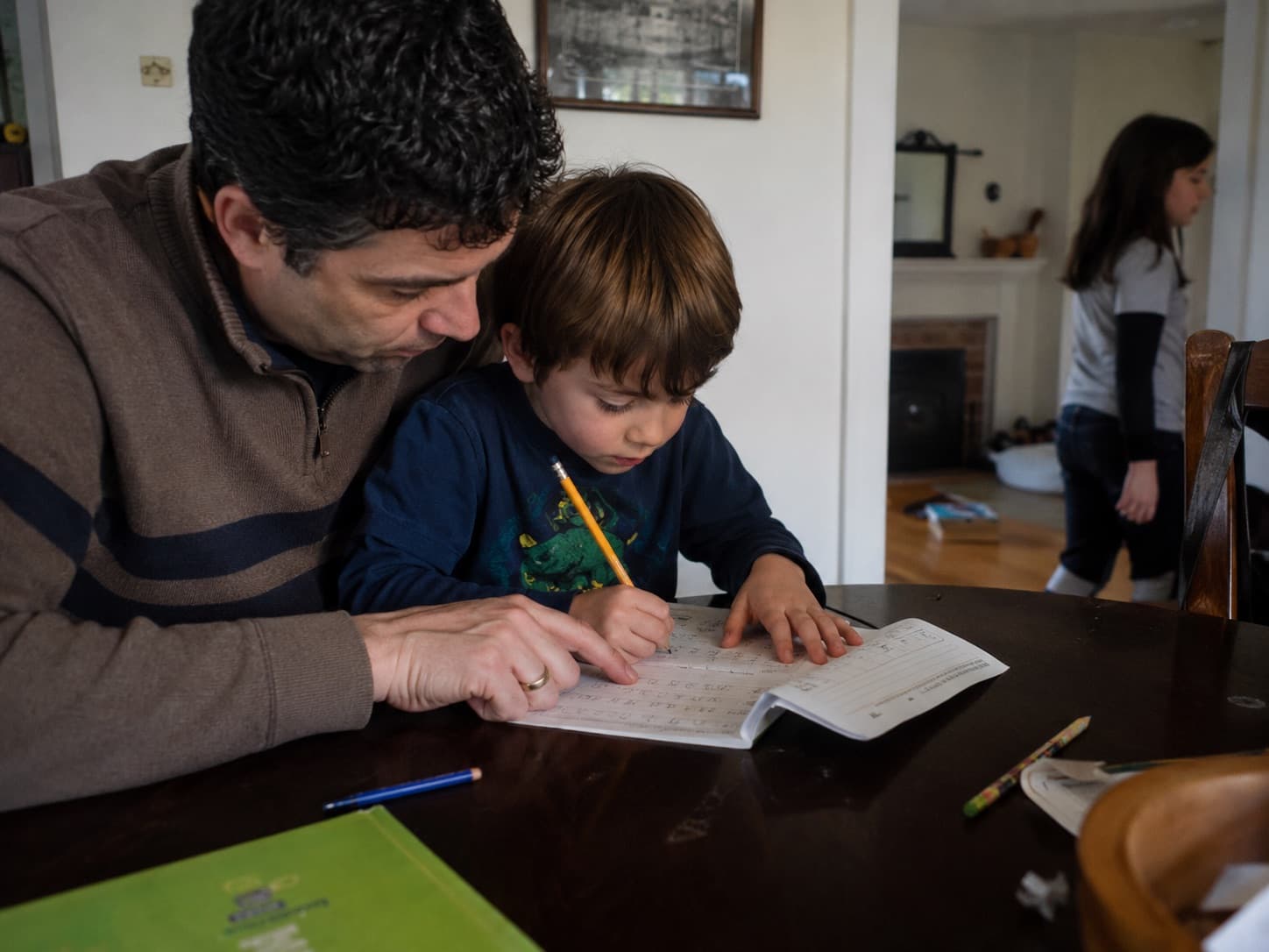 A father helping his son with handwriting practice at the dining table.