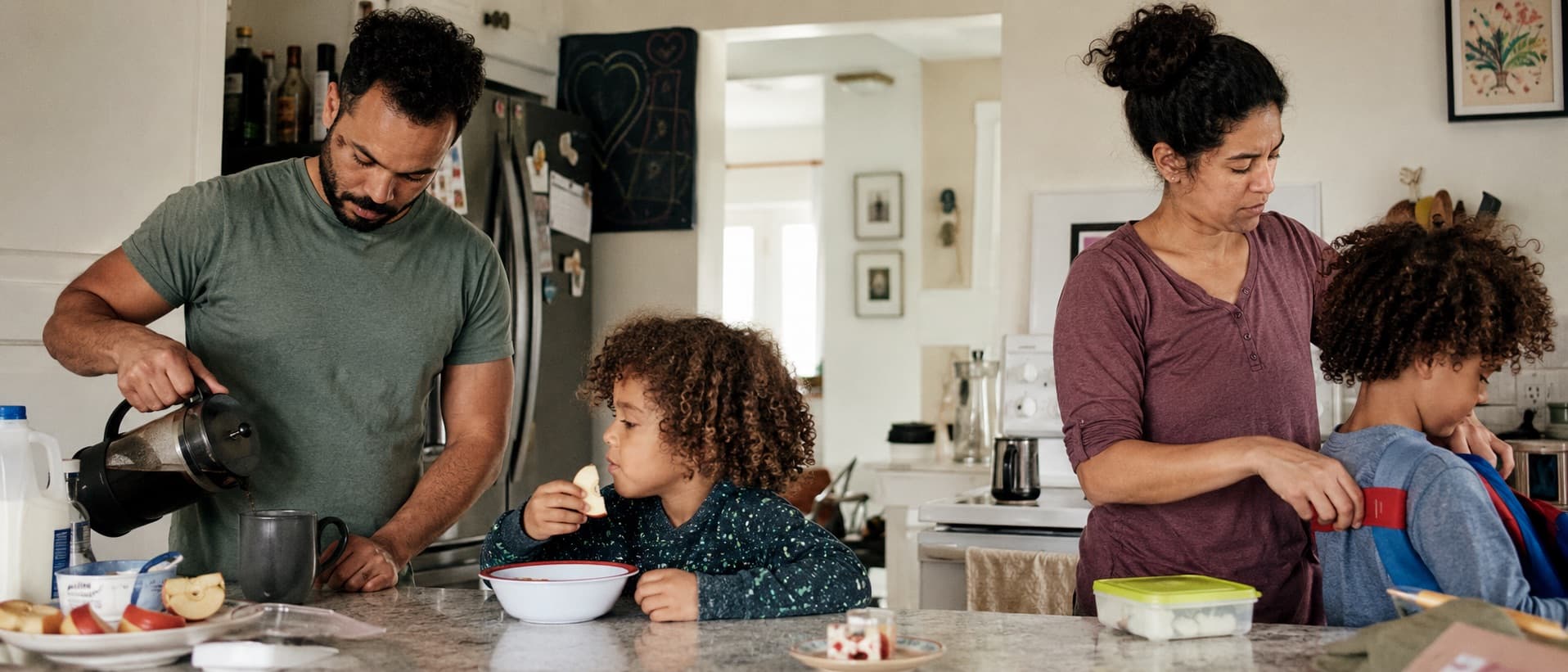 A family at breakfast before school: coffee poured, an apple, a backpack being fastened.